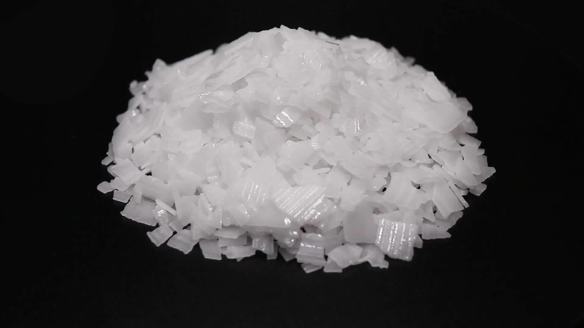 A pile of translucent white crystalline flakes is displayed against a black background. The flakes vary slightly in size and shape, with some appearing more angular and others more irregular. The texture suggests a solid but brittle material.