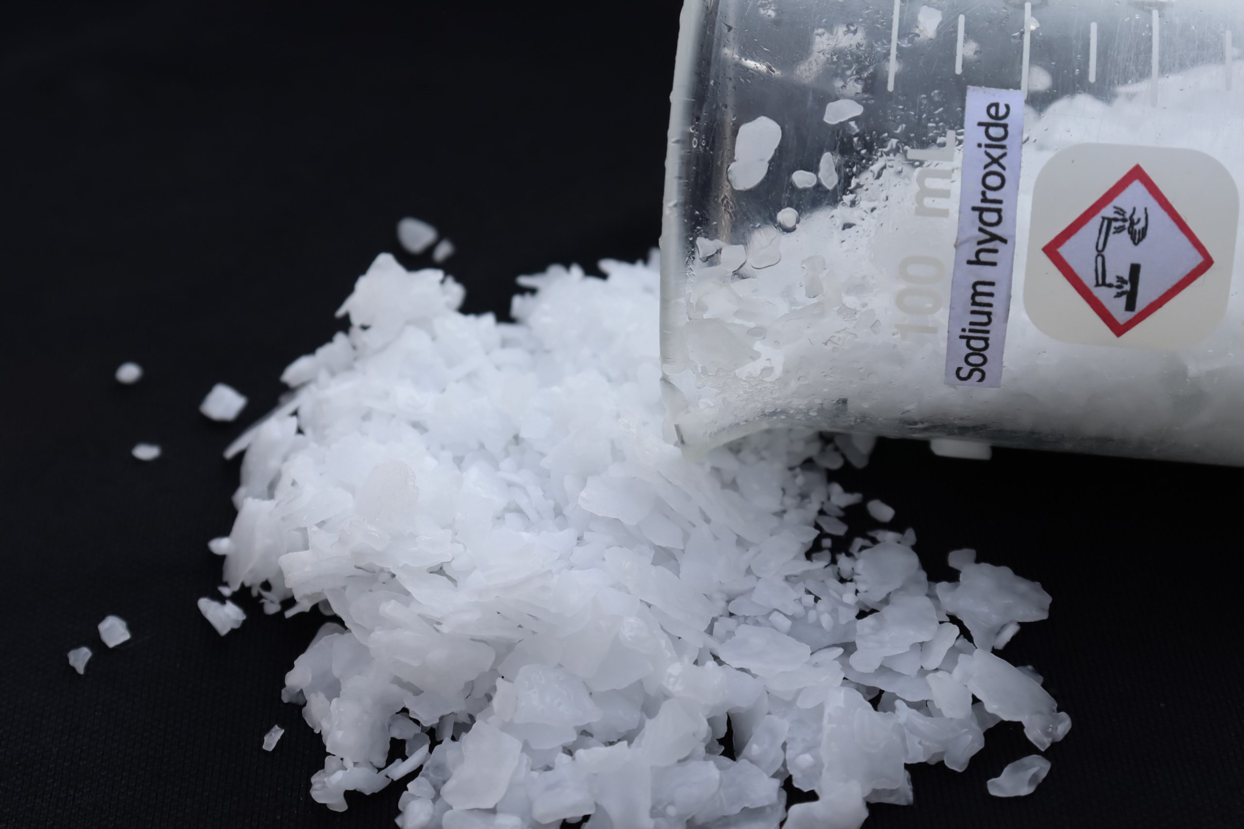 A beaker labeled "Sodium Hydroxide" is tipped on its side, spilling white crystalline flakes onto a black surface.