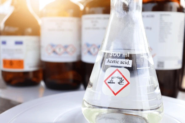 Close-up of a glass flask containing acetic acid, labeled with a hazardous material warning. The flask is placed on a white surface. In the blurred background, there are several brown bottles with various labels and hazard symbols.