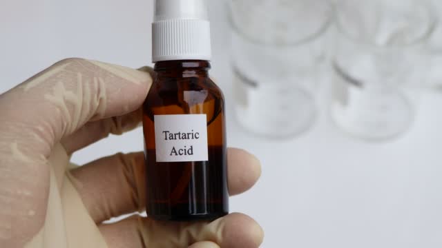 A gloved hand holding a small brown bottle labeled "Tartaric Acid." The bottle has a white cap, and the background shows several blurred glass containers.