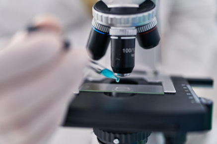 Close-up shot of a microscope's objective lenses focusing on a slide. The background is blurred in light blue, highlighting the microscope's metal and glass components. The image emphasizes the precision and detail of the scientific equipment.