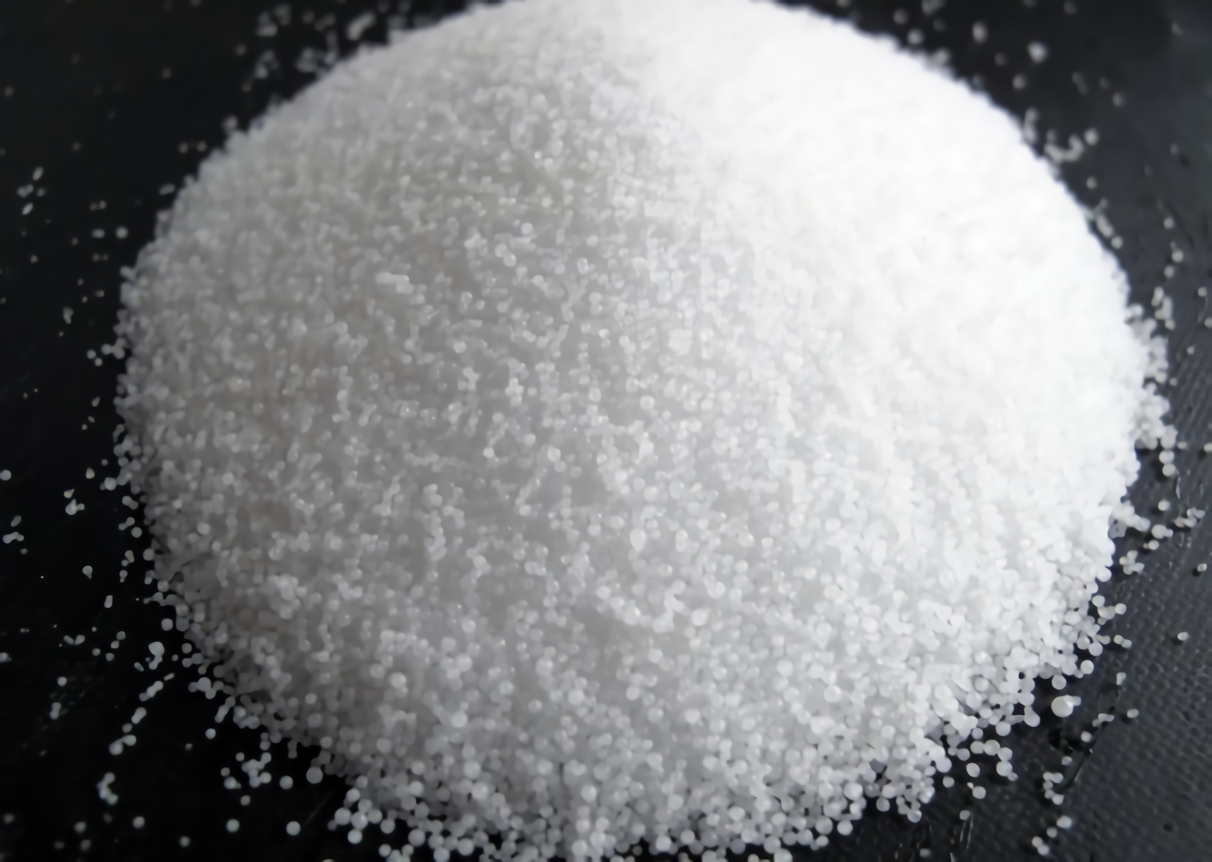 A close-up photo of a mound of coarse, white granules resembling salt or sugar on a black surface. The granules appear uniform in size and shape, giving the mound a smooth, even texture.