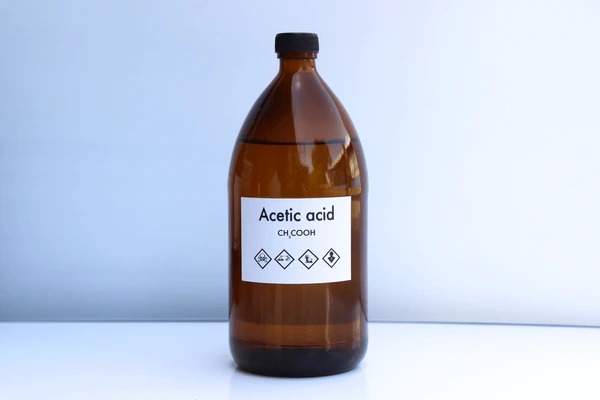 A brown glass bottle labeled "Acetic acid CH₃COOH" sits on a white surface against a light background. The label includes four hazard symbols indicating caution, flammable, irritant, and corrosive.