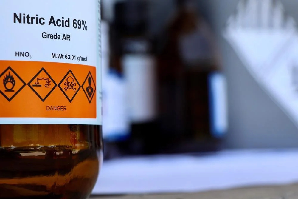 Close-up of a bottle labeled "Nitric Acid 69%" with hazard symbols, focused in the foreground against a blurred laboratory backdrop.