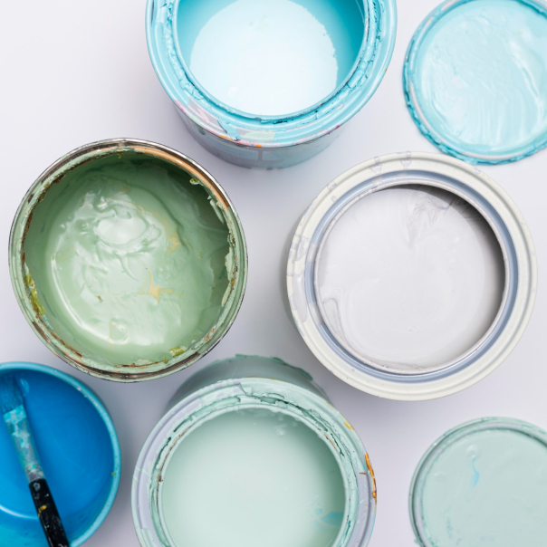 Open paint cans in various shades of blue and green, viewed from above, with a paintbrush visible. The paint cans show signs of use with drips along the edges.