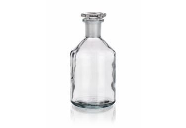 A clear glass bottle with a round base and a narrow neck, topped with a matching clear glass stopper. The bottle is empty and stands on a white reflective surface.