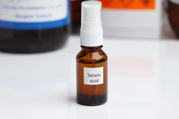 A small amber bottle labeled "Tartaric Acid" with a white spray pump top is centered in the image, against a white background. Other blurred chemical containers are visible in the background.