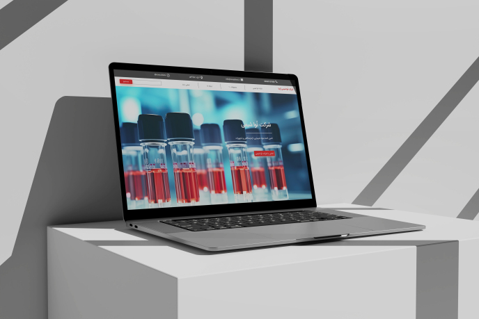 A sleek laptop open on a white, angular stand displaying a high-resolution image of test tubes filled with red liquid on its screen. the background features geometric gray and white shapes.